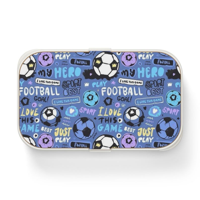 May include: A rectangular lunch box with a blue background featuring a repeating pattern of soccer balls, purple circles, and text including "MY HERO," "FOOTBALL," "GOAL," "I LOVE THIS GAME," "SPORT," "BEST," and "JUST PLAY" in white and light blue.