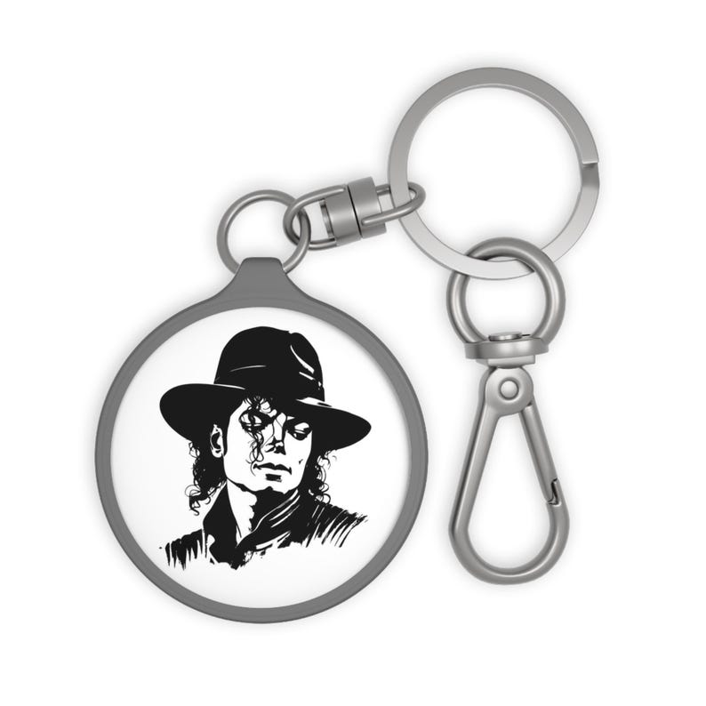 May include: A round keychain featuring a black and white illustration of a person wearing a fedora hat. The keychain has a silver-colored ring and clasp for attaching to keys or bags. The image is a portrait.