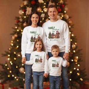 K&ouml;nnte beinhalten: Wei&szlig;e lang&auml;rmelige Rundhals-Sweatshirts mit einer Grafik von vier Dackeln in festlicher Kleidung, einem Weihnachtsbaum und dem Text "FAMILY Christmas 2026... The Williams". Die Sweatshirts werden von zwei Erwachsenen und zwei Kindern mit blauen Jeans getragen.