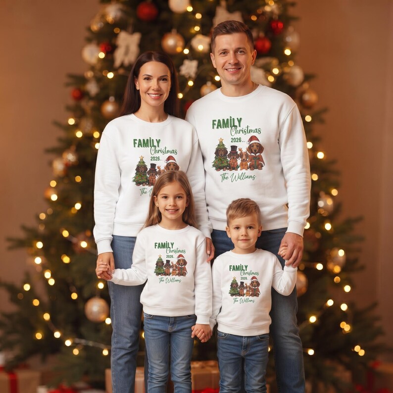 K&ouml;nnte beinhalten: Wei&szlig;e lang&auml;rmelige Rundhals-Sweatshirts mit einer Grafik von vier Dackeln in festlicher Kleidung, einem Weihnachtsbaum und dem Text "FAMILY Christmas 2026... The Williams". Die Sweatshirts werden von zwei Erwachsenen und zwei Kindern mit blauen Jeans getragen.