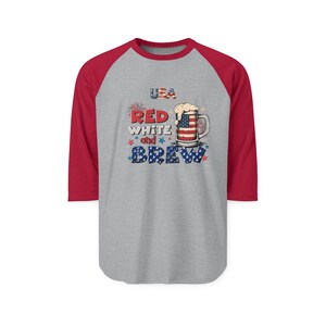 Peut inclure: Un t-shirt de baseball gris avec des manches trois-quarts rouges pr&eacute;sentant un graphique d'une chope de bi&egrave;re avec un motif de drapeau am&eacute;ricain, de la mousse et le texte USA RED WHITE and BREW avec des petites &eacute;toiles rouges et bleues.
