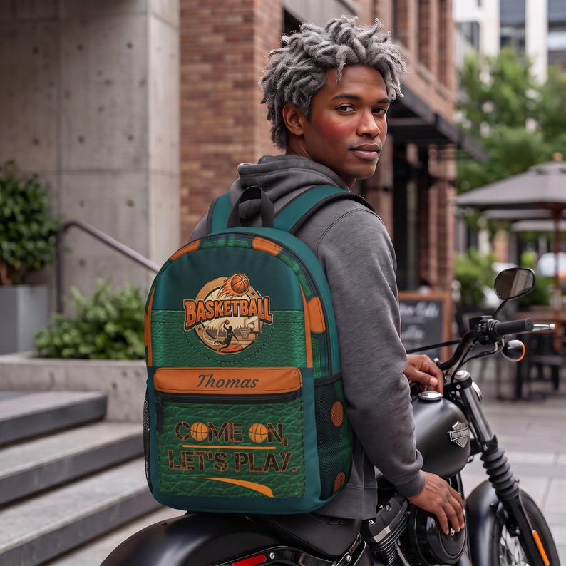 May include: A dark green backpack featuring a basketball graphic with the text "BASKETBALL" and "COME ON, LET'S PLAY." The bag includes a brown horizontal accent strip with the name "Thomas" printed in a script font and a textured surface.