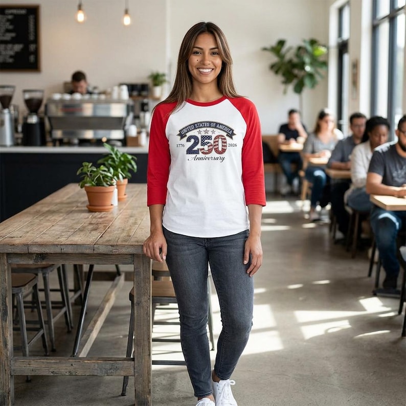 May include: A white and red baseball tee with the text "UNITED STATES OF AMERICA 250th Anniversary 1776-2026". The shirt is worn by a person in a cafe setting. The shirt has red sleeves and a white body.