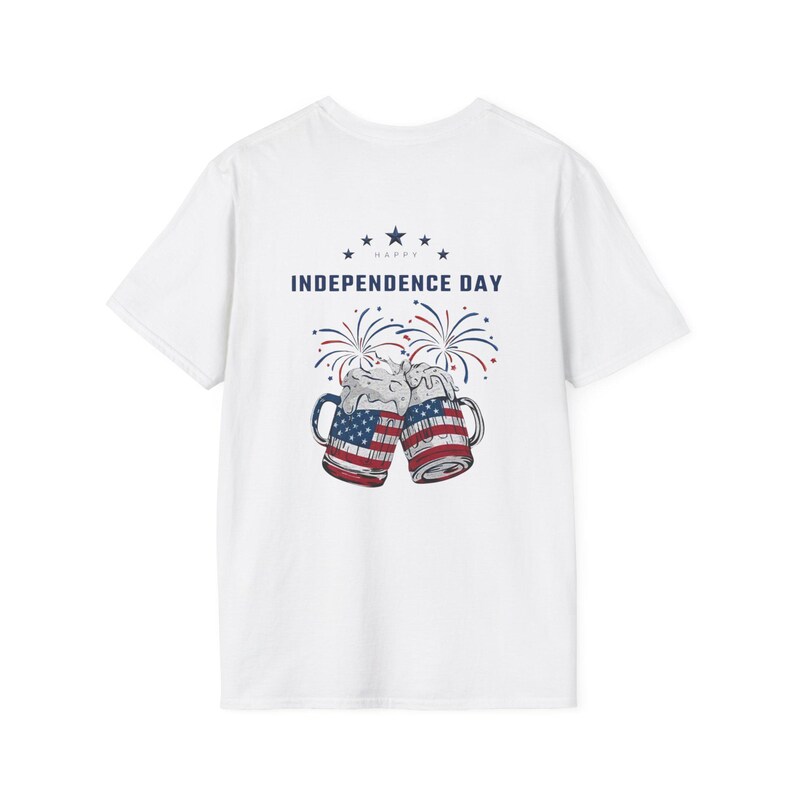 May include: A white t-shirt featuring a graphic of two beer mugs clinking together, each decorated with the American flag pattern. Above the mugs, the text reads HAPPY INDEPENDENCE DAY in blue, accompanied by red and blue firework illustrations.