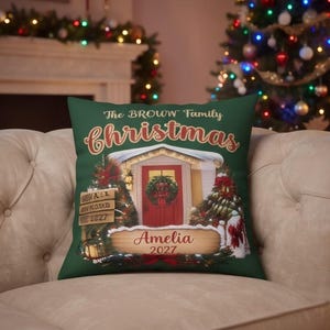 May include: A dark green square throw pillow featuring a printed illustration of a house entrance with a red door, a wreath, and Christmas trees. The text reads The BROWN Family christmas, Amelia, and 2027 on wooden-style signs.