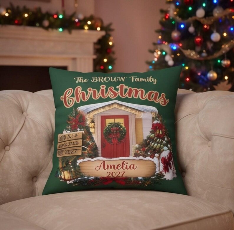 May include: A dark green square throw pillow featuring a printed illustration of a house entrance with a red door, a wreath, and Christmas trees. The text reads The BROWN Family christmas, Amelia, and 2027 on wooden-style signs.