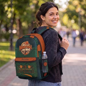May include: A dark green and orange backpack featuring a basketball graphic, the text "BASKETBALL", the name "Jessica", and the phrase "COME ON, LET'S PLAY." The bag has a side mesh pocket containing a clear plastic water bottle with a white cap.