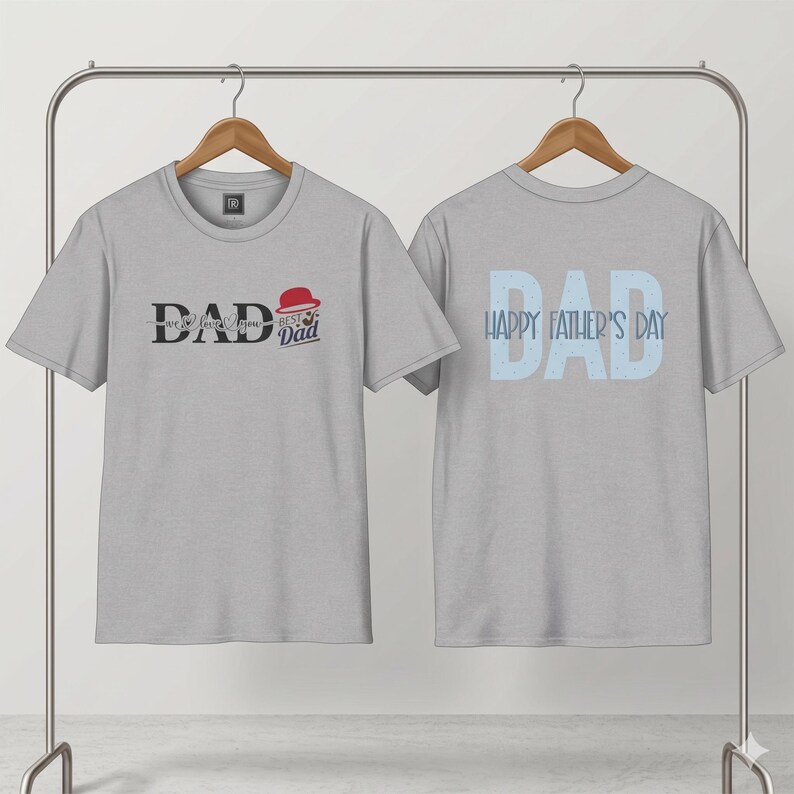 May include: Two light gray t-shirts hanging on a metal rack. The front of the first shirt has the word "DAD" with "we love you" below it, and a graphic of a hat. The second shirt has "DAD" and "HAPPY FATHER'S DAY" in light blue.