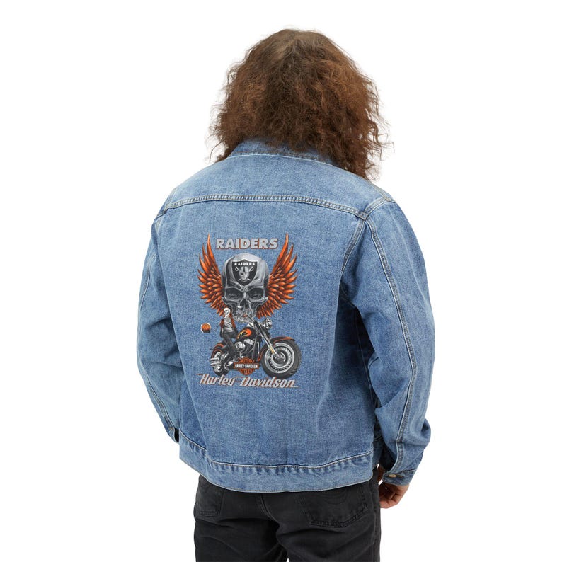May include: A light blue denim jacket featuring a graphic on the back with a skull wearing a Raiders helmet, large orange wings, and a motorcycle. The text reads Raiders and Harley Davidson in a light-colored font against the blue fabric.
