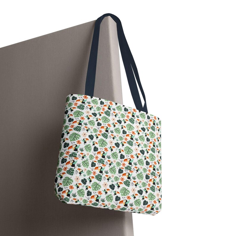 May include: A white tote bag with a repeating pattern of toucans, green leaves, and flowers. The bag has navy blue handles and is hanging against a neutral-colored wall. The design features orange, black, and pink accents.