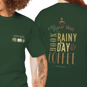 May include: A dark green t-shirt featuring a small graphic on the front left chest with a stack of books, an open book, and a coffee mug with the text "BOOK NERD". The back displays "Master blend: BOOK RAINY DAY & COFFEE" with a coffee cup icon.