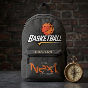 Może przedstawiać: Ciemnoszary plecak z grafiką piłki do koszyk&oacute;wki nad napisem "BASKETBALL" i "-CUSTOM-". Poniżej znajduje się napis "WE ARE Next Generations" w kolorze pomarańczowym i białym. Po prawej stronie plecaka znajduje się złoty kompas.