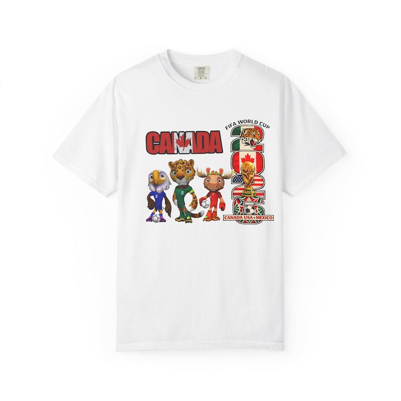 May include: White t-shirt featuring the word "CANADA" in red block letters, with the FIFA World Cup 2026 logo and mascot illustrations. The shirt includes the text "CANADA USA MEXICO".