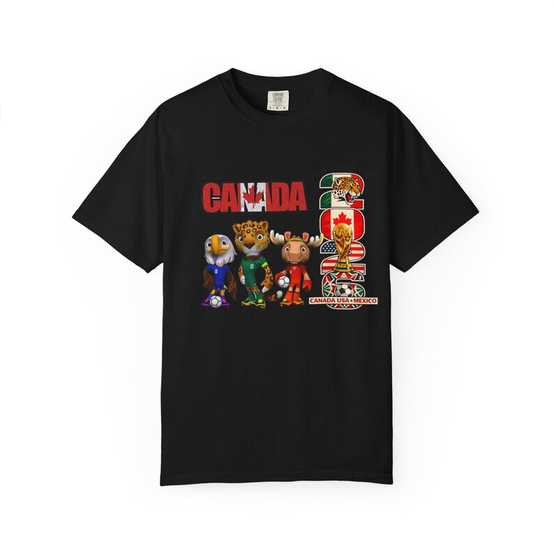 May include: Black t-shirt featuring the word "CANADA" in red, with the year "2026" and a graphic of the FIFA World Cup trophy. The shirt also displays cartoon animal mascots and the flags of Canada, Mexico, and the USA.
