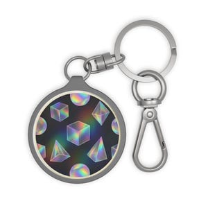 May include: A round keychain with a geometric design. The keychain features a black background with holographic cubes, spheres, and pyramids. The keychain has a silver-colored metal ring and clasp.