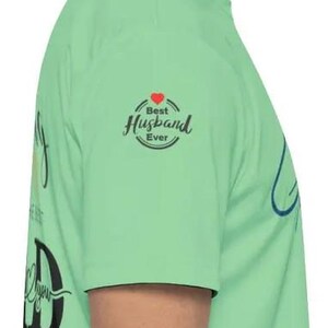 Pu&ograve; includere: Maglietta verde chiaro con la scritta "Best Husband Ever" in un design circolare con un cuore rosso. La maglietta presenta anche la parola "Dad" in un font grande.