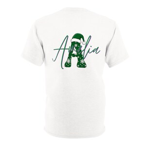Puede incluir: Una camiseta blanca de manga corta con un gr&aacute;fico en la espalda. El dise&ntilde;o muestra una letra A grande de color verde con copos de nieve blancos, coronada por un gorro de Pap&aacute; Noel verde, y el nombre Amelia escrito en cursiva encima.