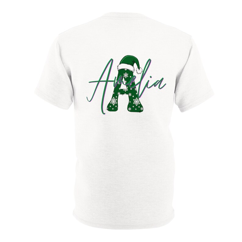 Puede incluir: Una camiseta blanca de manga corta con un gr&aacute;fico en la espalda. El dise&ntilde;o muestra una letra A grande de color verde con copos de nieve blancos, coronada por un gorro de Pap&aacute; Noel verde, y el nombre Amelia escrito en cursiva encima.