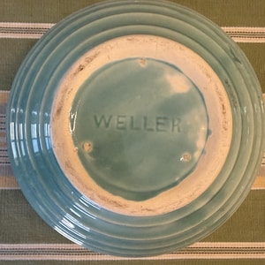 May include: A round, light blue ceramic bowl with a ribbed exterior design. The underside features a circular base with the word WELLER embossed in the center. The glaze is a consistent light blue color, and the base shows a white ceramic rim.