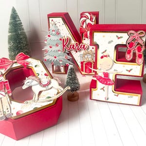 May include: Red and gold Christmas-themed decorative boxes and letters. The letters spell out "KARA" and feature a Christmas tree, a ballerina, a rocking horse, and a nutcracker. The letters are decorated with gold trim and festive accents.