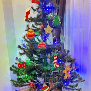 May include: A green artificial Christmas tree decorated with colorful knitted ornaments, including a stocking, a star, a gingerbread man, a candy cane, a Christmas tree, and ornaments shaped like balls. The tree is lit with white and blue fairy lights.
