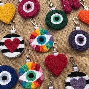 Puede incluir: Surtido de llaveros de crochet coloridos con varios diseños, incluyendo corazones, ojos malos y letras. Los llaveros están hechos con hilo de lana en tonos de rojo, azul, verde, amarillo, morado y blanco.