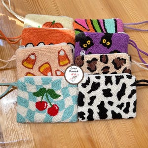 Puede incluir: Una colección de ocho estuches rectangulares con cremallera hechos con punch needle y correas de muñeca. Los diseños incluyen una calabaza, naranja sólido, maíz dulce, cerezas sobre cuadros azules, rayas, gatos negros, leopardo y vaca. Texto Love Punch Art.