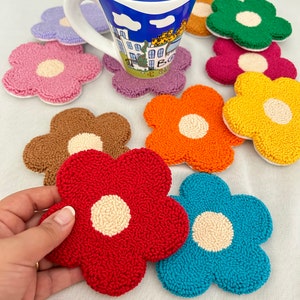 May include: A collection of colorful, flower-shaped coasters in various colors, including red, blue, pink, and yellow. Each coaster has a cream-colored center. A white mug with a blue and white design sits atop one of the coasters. The coasters are made of a textured material.