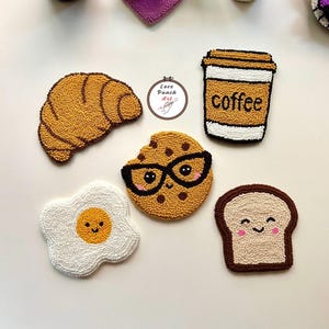 May include: A selection of handmade, textured coasters with breakfast and coffee designs. Includes a croissant, a cookie wearing glasses, a fried egg, a coffee cup, and toast. The coasters are in shades of brown, white, and yellow.