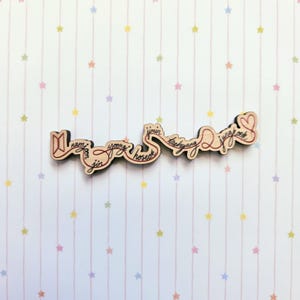 May include: A light-colored wooden cutout featuring the BTS logo on the left and a heart on the right, connected by a continuous flowing line of cursive text that spells out the names Namjoon, Jin, Yoongi, Hoseok, Jimin, Taehyung, and Jungkook.