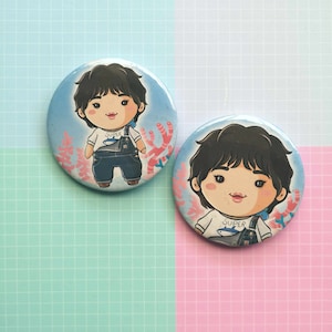 Op de afbeelding: Twee ronde buttons met een cartoonillustratie van een persoon met zwart haar, gekleed in een wit shirt met een haaienafbeelding en een blauwe overall. Het woord "SUPER" staat op het shirt. De achtergrond is lichtblauw met koraal.