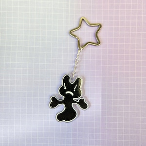 Puede incluir: Un llavero en blanco y negro con un personaje de dibujos animados con una cara triste. El llavero tiene un anillo plateado en forma de estrella y una cadena. El fondo es una cuadrícula morada clara.