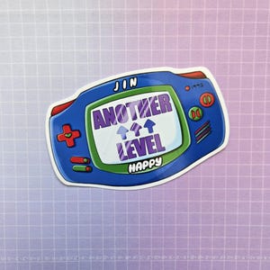 May include: A blue and green game console sticker with the text "JIN ANOTHER LEVEL HAPPY" and three upward arrows on the screen.