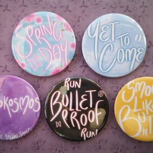 Op de afbeelding: Vijf ronde knoppen met verschillende ontwerpen en tekst. De knoppen zijn paars, blauw, zwart, geel en lichtblauw. De tekst op de knoppen luidt "Mikrokosmos", "Yet To Come", "Run Bulletproof Run", "Smooth Like Butter" en "Spring Day".