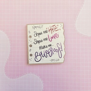 Puede incluir: Una pegatina rectangular con un borde rasgado, con texto manuscrito en negro, morado y rosa. El texto incluye frases como "Show me hate" y "Show me love", con "Bulletproof!" en letra grande. Fondo rosa cuadriculado.