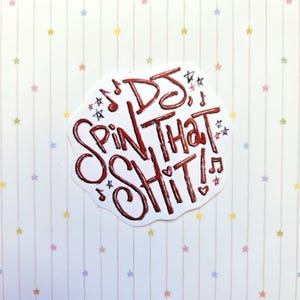 以下が含まれることがあります： 赤の手書き風テキスト「DJ SPIN THAT SHIT!」が描かれた白いダイカットステッカー。スタイリッシュで質感のあるフォントで、文字の周りには小さな赤と黒の音符、星、ハートのモチーフが散りばめられています。