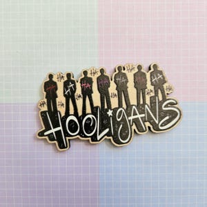 May include: A wooden pin featuring seven black silhouettes of figures standing in a row. Above the figures, the word HA is repeated multiple times in black and pink. Below the figures, the word HOOLIGANS is written in large white script.
