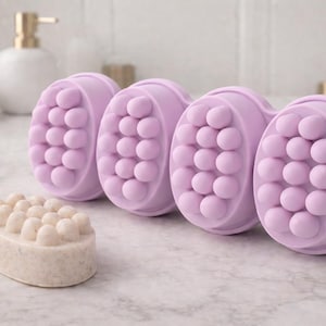 Könnte beinhalten: Vier lilafarbene Massage-Tools mit abgerundeten, strukturierten Oberflächen sind in einer Reihe angeordnet. Eine hellbeige ovale Seife mit ähnlicher strukturierter Oberfläche liegt davor. Die Artikel befinden sich auf einer Marmoroberfläche, was auf ein Spa- oder Self-Care-Thema hindeutet.