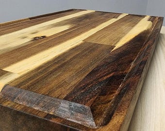 May include: A rectangular wooden cutting board with a dark brown and light tan wood grain pattern. The board has a juice groove around the edge and is made of multiple wood pieces. The board is on a light wood surface.