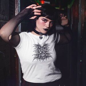May include: A person wearing a white t-shirt with a black graphic design that says "Death Metal" in a gothic font. They are wearing black fishnet gloves and a black choker with a silver heart pendant.