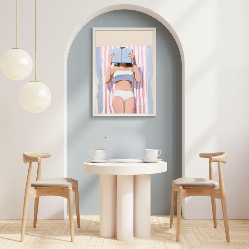 May include: A woman in a white swimsuit is lying on a pink and white striped towel, reading a blue book. The scene is set in a minimalist living room with a white table and two chairs.