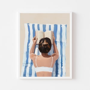 Op de afbeelding: Een ingelijste kunstdruk toont een persoon die een boek leest op het strand. De persoon draagt een witte top en ligt op een blauw-wit gestreepte handdoek. De achtergrond is lichtbeige, wat duidt op een strandscène.