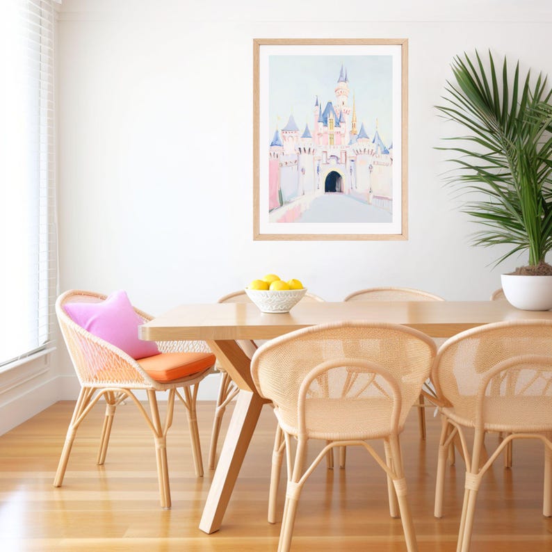 May include: A dining room scene featuring a light wood table and woven chairs with colorful cushions. A framed watercolor painting of a castle hangs on the white wall. A bowl of lemons sits on the table, and a potted plant is in the corner.