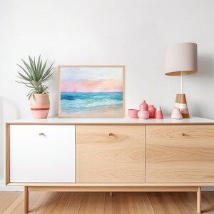 Peut inclure: Une peinture &agrave; l'aquarelle encadr&eacute;e d'un paysage marin avec un ciel rose et bleu, une eau turquoise et des vagues blanches qui se brisent sur une plage de sable.