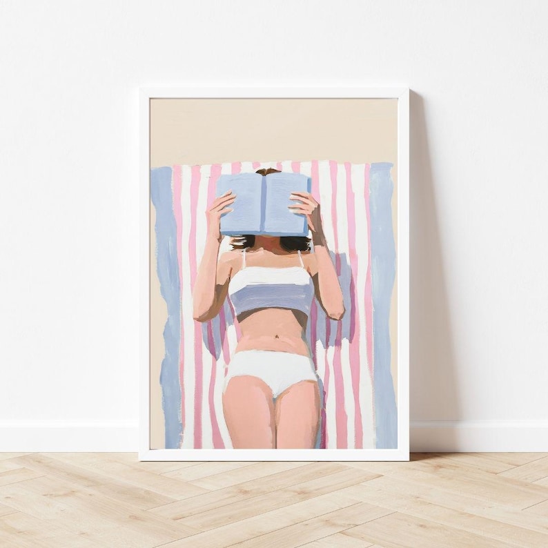 May include: A woman in a white bikini lies on a pink and blue striped beach towel, reading a blue book.