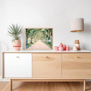 K&ouml;nnte beinhalten: Aquarellmalerei eines von B&auml;umen ges&auml;umten Weges mit einer rosafarbenen Stra&szlig;e. Das Kunstwerk befindet sich in einem hellen Holzrahmen. Das Gem&auml;lde wird auf einem wei&szlig;en und hellen Holzschrank neben einer Topfpflanze und einer Lampe ausgestellt.
