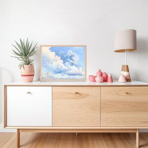 Puede incluir: Una pintura enmarcada de un cielo azul con nubes blancas. La pintura est&aacute; colgada en una pared blanca sobre una c&oacute;moda de madera clara con tres cajones. Hay jarrones de cer&aacute;mica rosa y blanca sobre la c&oacute;moda.