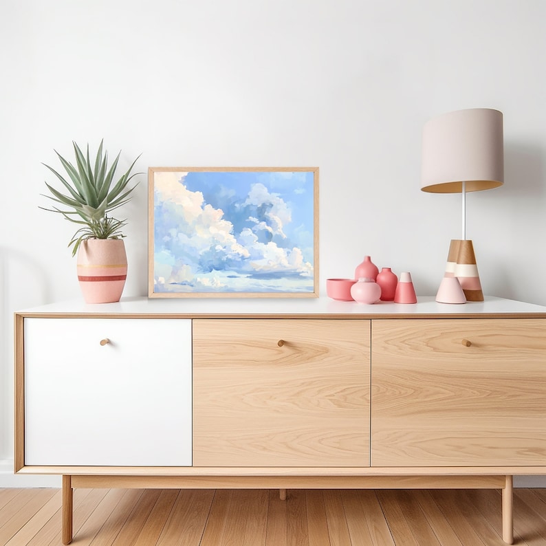 Puede incluir: Una pintura enmarcada de un cielo azul con nubes blancas. La pintura est&aacute; colgada en una pared blanca sobre una c&oacute;moda de madera clara con tres cajones. Hay jarrones de cer&aacute;mica rosa y blanca sobre la c&oacute;moda.