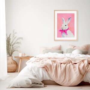 May include: A white rabbit with a pink bow sits on a pink background in a framed print. The print is hanging on a white wall above a bed with white and pink bedding.