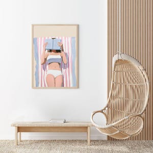 May include: A framed illustration of a person lying on a striped beach towel, reading a book. The person is wearing a white bikini and has their face obscured by the book. The illustration is in a minimalist style with muted colors.